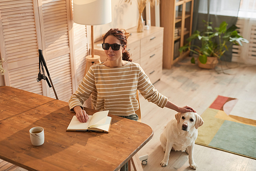 Warm toned portrait of modern blind woman reading braille book while sitting at table in cozy home interior and petting guide dog, copy space