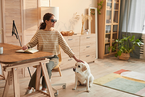 Warm toned full length portrait of modern blind woman sitting at table in cozy home interior and petting guide dog, copy space