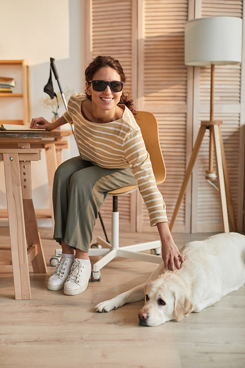 Vertical full length portrait of modern blind woman sitting at table in cozy home interior and petting guide dog