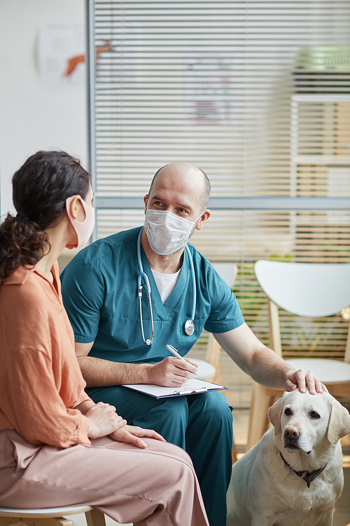 Vertical portrait of young woman wearing mask while talking to veterinarian in waiting room at vet clinic with white dog
