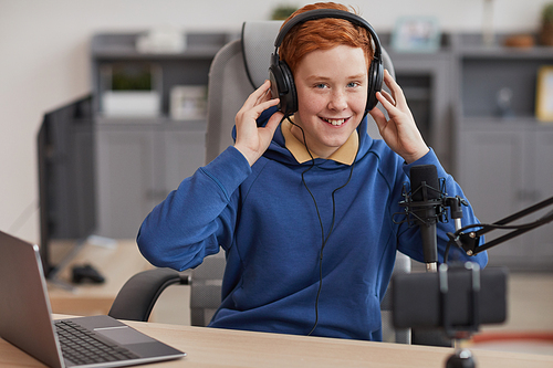 Portrait of red haired teenage boy wearing headphones and looking at camera while filming videos at home, copy space
