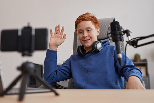 Portrait of red haired teenage boy waving to camera while recording videos or online streaming at home, copy space