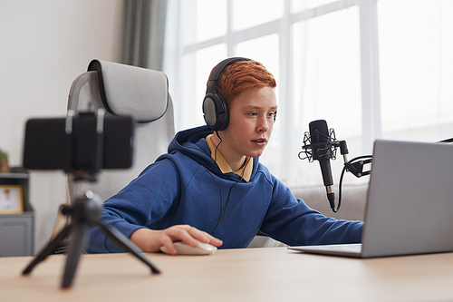 Portrait of red haired teenage boy playing video games with microphone and camera set up for online streaming, copy space