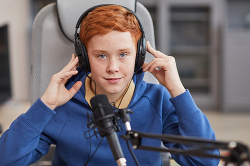 Front view portrait of red haired teenage boy speaking to microphone and wearing headphones while recording podcast or online streaming