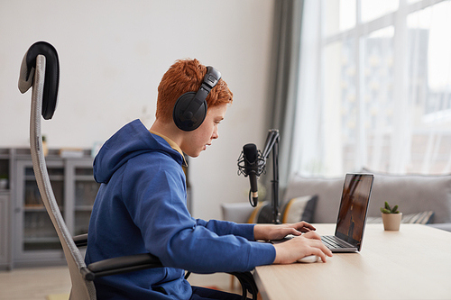 Side view portrait of red haired teenage boy playing video games with headphones and microphone set up for online streaming, copy space