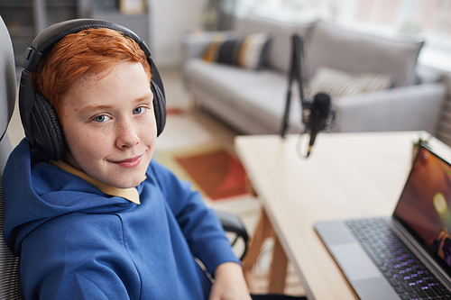 High angle portrait of smiling red haired boy looking at camera while playing video games with microphone set up for online streaming, copy space