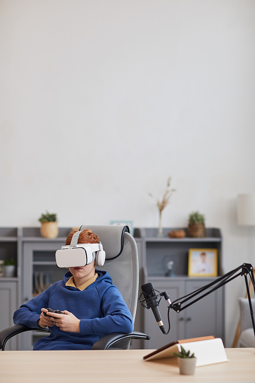 Vertical wide angle portrait of teenage boy wearing VR headset and holding gamepad while playing immersive video games at home, copy space