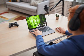 Back view portrait of red haired teenage boy playing video games via laptop with microphone stand for online streaming, copy space