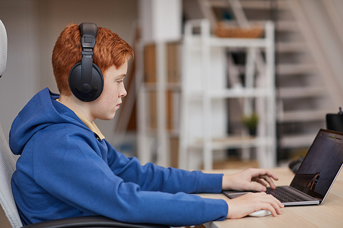 Side view portrait of red haired teenage boy playing video games via laptop with camera set up for online streaming, copy space