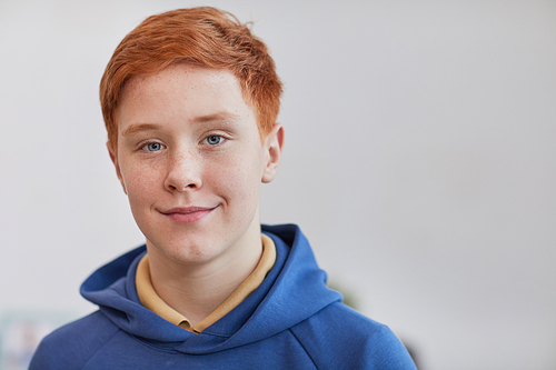 Head and shoulders portrait of freckled red haired boy looking at camera and smiling while standing against white background, copy space