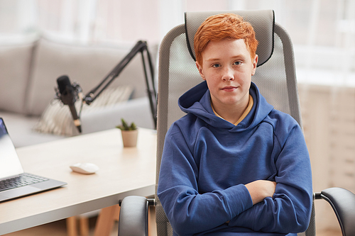 Portrait of red haired teenage boy looking at camera while sitting in chair against computer in background, copy space