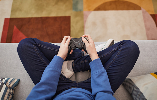 Top down view at unrecognizable teenage boy playing video games while sitting on sofa and holding gamepad, copy space