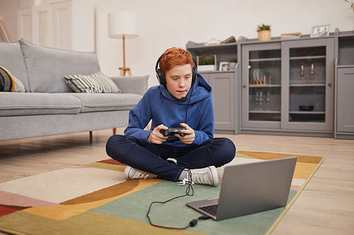 Full length portrait of red haired teenage boy playing video games while sitting on floor and holding gamepad, copy space