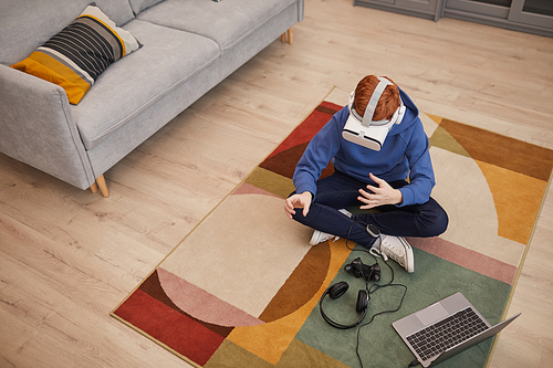 Top down view at red haired teenage boy playing video games in VR while sitting on floor on graphic carpet, copy space