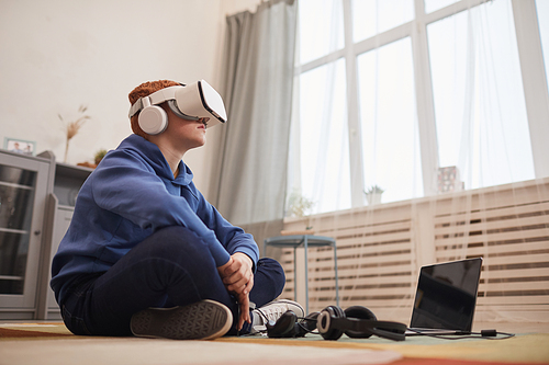 Side view portrait o red haired teenage boy wearing VR headset while playing video games sitting on floor, copy space