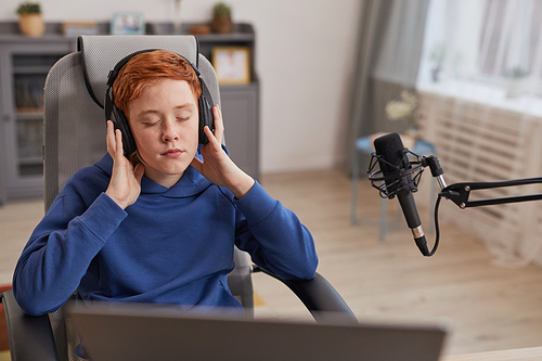 Portrait of red haired teenage boy wearing headphones and enjoying music with eyes closed while relaxing or recording podcast, copy space