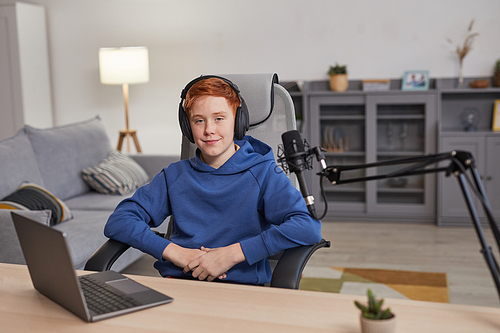 Portrait of red haired teenage boy wearing headphones and smiling at camera while sitting at desk with microphone set up for podcasting or online streaming, copy space