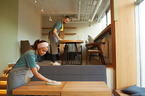 Side view portrait of two young waiters cleaning tables in coffee shop with warm wooden accents, copy space