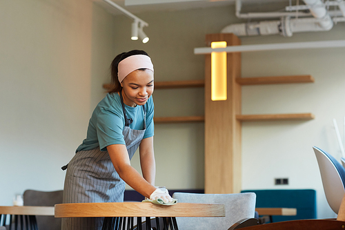Portrait of smiling mixed-race waitress cleaning table in cafe or coffee shop while preparing for opening in morning, copy space