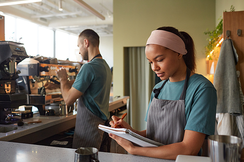 Waist up portrait of young African-American woman working in coffee shop and holding clipboard, copy space