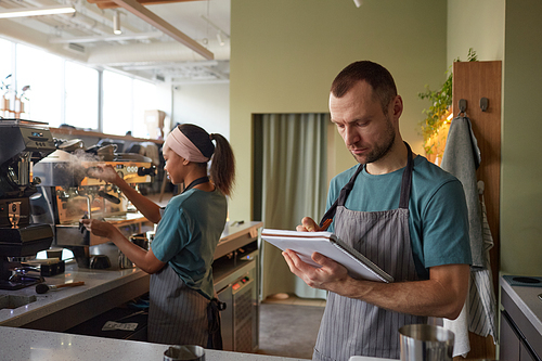 Waist up portrait of male barista wearing apron while working in coffee shop and holding clipboard, copy space