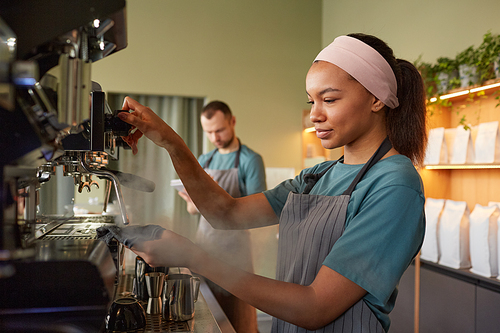 Side view portrait of young female barista making fresh coffee in cafe and operating coffee machine