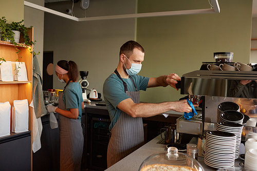 Side view portrait of male barista wearing mask and using coffee machine while making fresh coffee at bar in cafe, copy space
