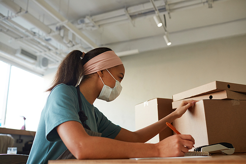 Side view portrait of young African-American woman wearing mask while unpacking boxes and doing inventory at small business shop, copy space