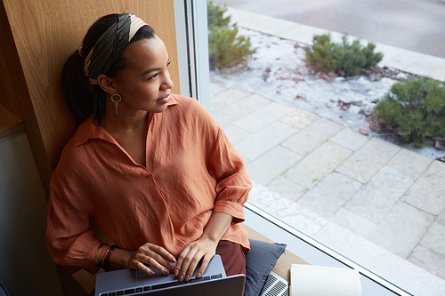 High angle portrait of young African-American businesswoman using laptop while looking at window in cafe and enjoying remote work, copy space