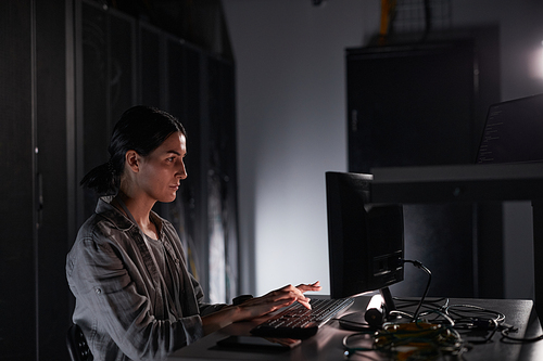 Side view portrait of female network engineer using laptop while sitting in dark server room, copy space