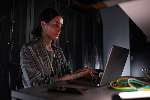 Portrait of female network engineer using laptop while sitting in dark server room, copy space