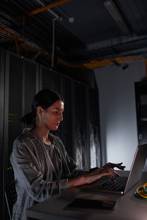 Vertical side view portrait of female network engineer using laptop while sitting in dark server room, copy space