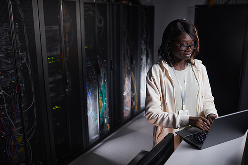 Portrait of African-American female network engineer using laptop while working in dark server room, copy space