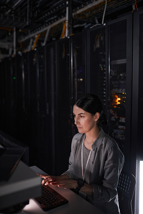 Vertical portrait of female network engineer using computer while working in dark server room, copy space