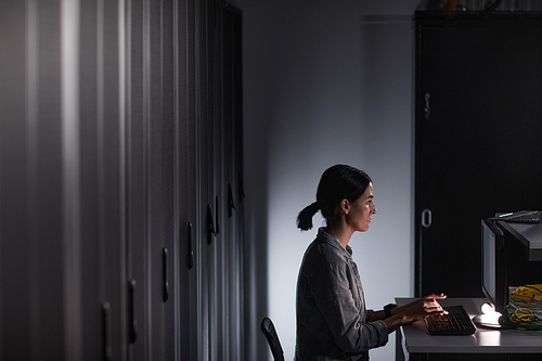 Graphic side view portrait of female network engineer using computer while working in dark server room, copy space