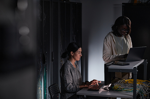 Portrait of two female IT engineers working with computer in dark server room, copy space