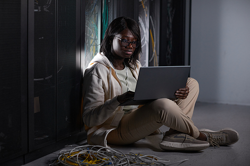 Full length portrait of young African-American woman using laptop in server room and sitting on floor while working with supercomputer at data center, copy space