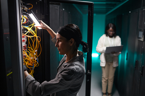Side view portrait of female network engineer connecting cables in server cabinet while working with supercomputer in data center, copy space
