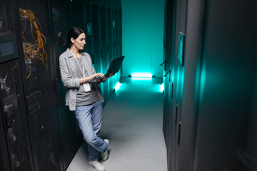 Full length high angle portrait of female data engineer holding laptop while working with supercomputer in server room, copy space
