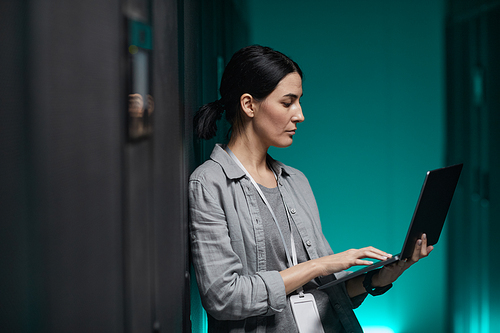 Side view portrait of female data engineer holding laptop while working with supercomputer in server room, copy space