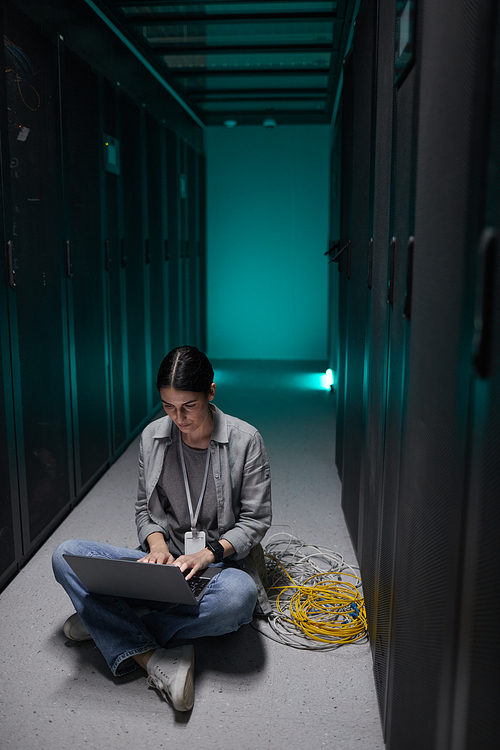Vertical portrait of female data engineer using laptop while sitting on floor in server room and setting up supercomputer network, copy space