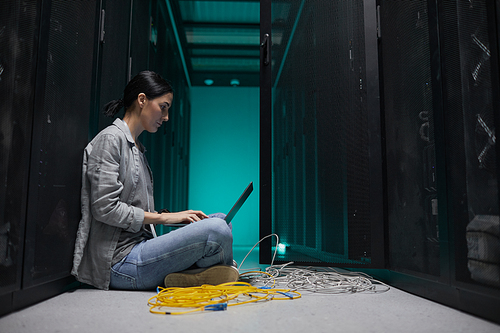 Side view portrait of female data engineer using laptop while sitting on floor in server room and setting up supercomputer network, copy space