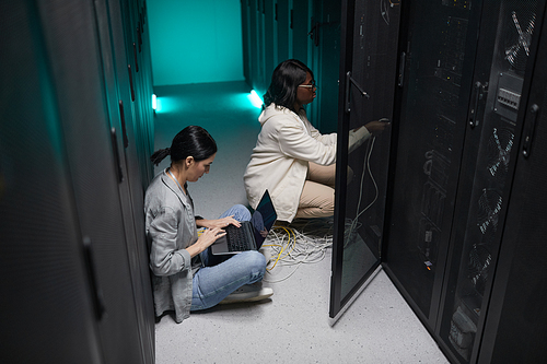 Portrait of two female data engineers using laptop in server room and setting up supercomputer network, copy space