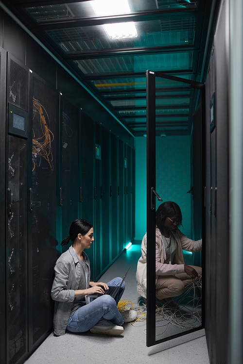 Vertical portrait of two female data engineers using laptop in server room and setting up supercomputer network, copy space