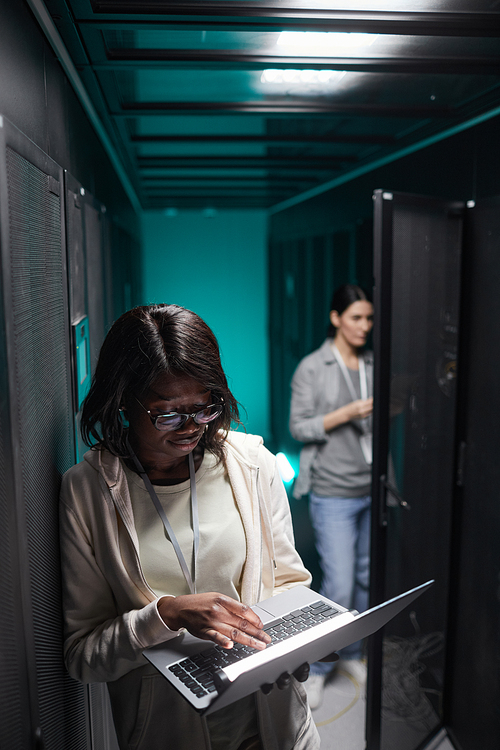 Vertical portrait of female data engineer using laptop in server room while setting up supercomputer network, copy space