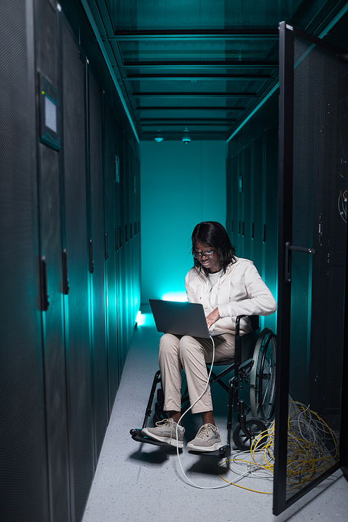Vertical full length portrait of disabled African-American woman using laptop while working in server room, accessible job opportunity
