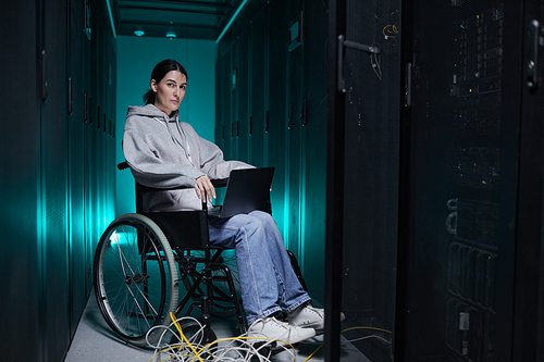 Full length portrait of disabled woman looking at camera while using laptop in server room, accessible job concept, copy space