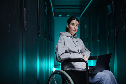 Portrait of disabled woman in wheelchair looking at camera while working with supercomputer network in server room, accessible job concept, copy space