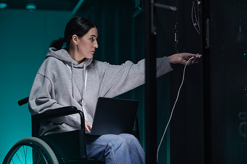Side view portrait of disabled woman in wheelchair working with supercomputer network in server room, accessible job concept, copy space