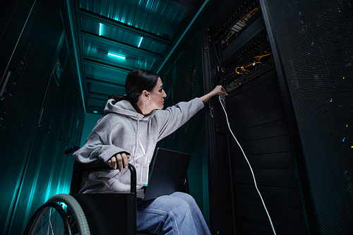 Low angle portrait of disabled woman in wheelchair working with supercomputer network in server room, accessible job concept, copy space